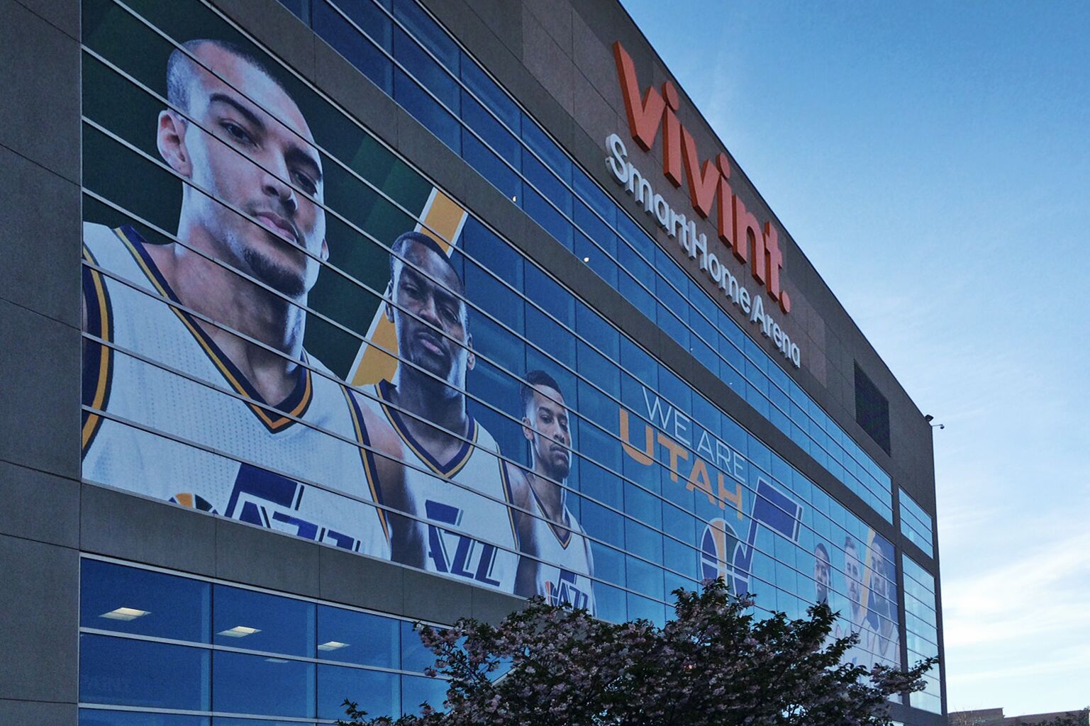 Außenansicht der Arena der Utah Jazz. Auf der Glasfassade befindet sich ein großes Wandbild mit mehreren Basketballspielern des Teams und dem Slogan 'We are Utah'.
