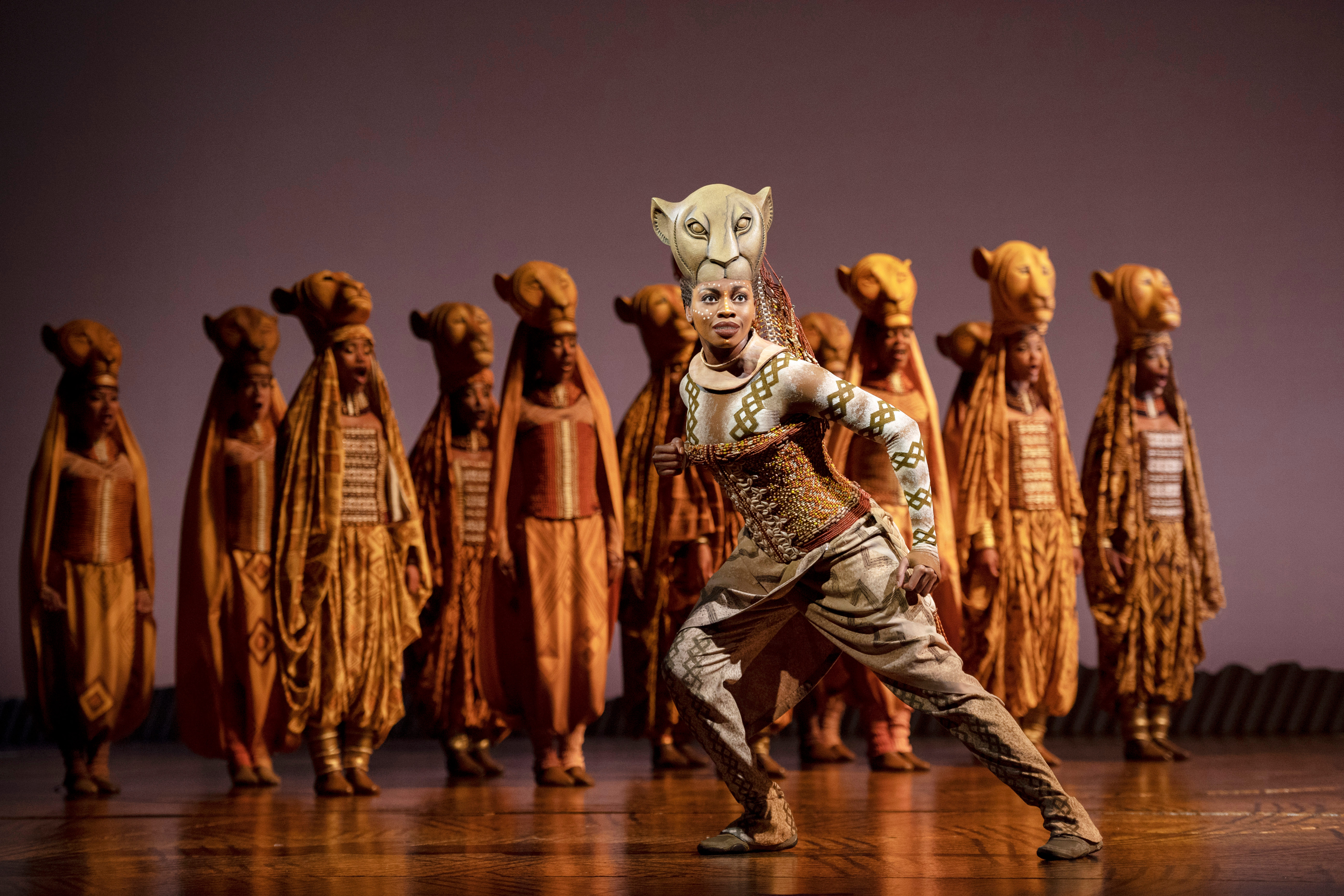 Szene aus dem Musical Der König der Löwen: Eine Darstellerin im Löwenkostüm steht im Vordergrund, während eine Gruppe von Tänzern in afrikanisch inspirierten Kostümen im Hintergrund eine Formation bildet.