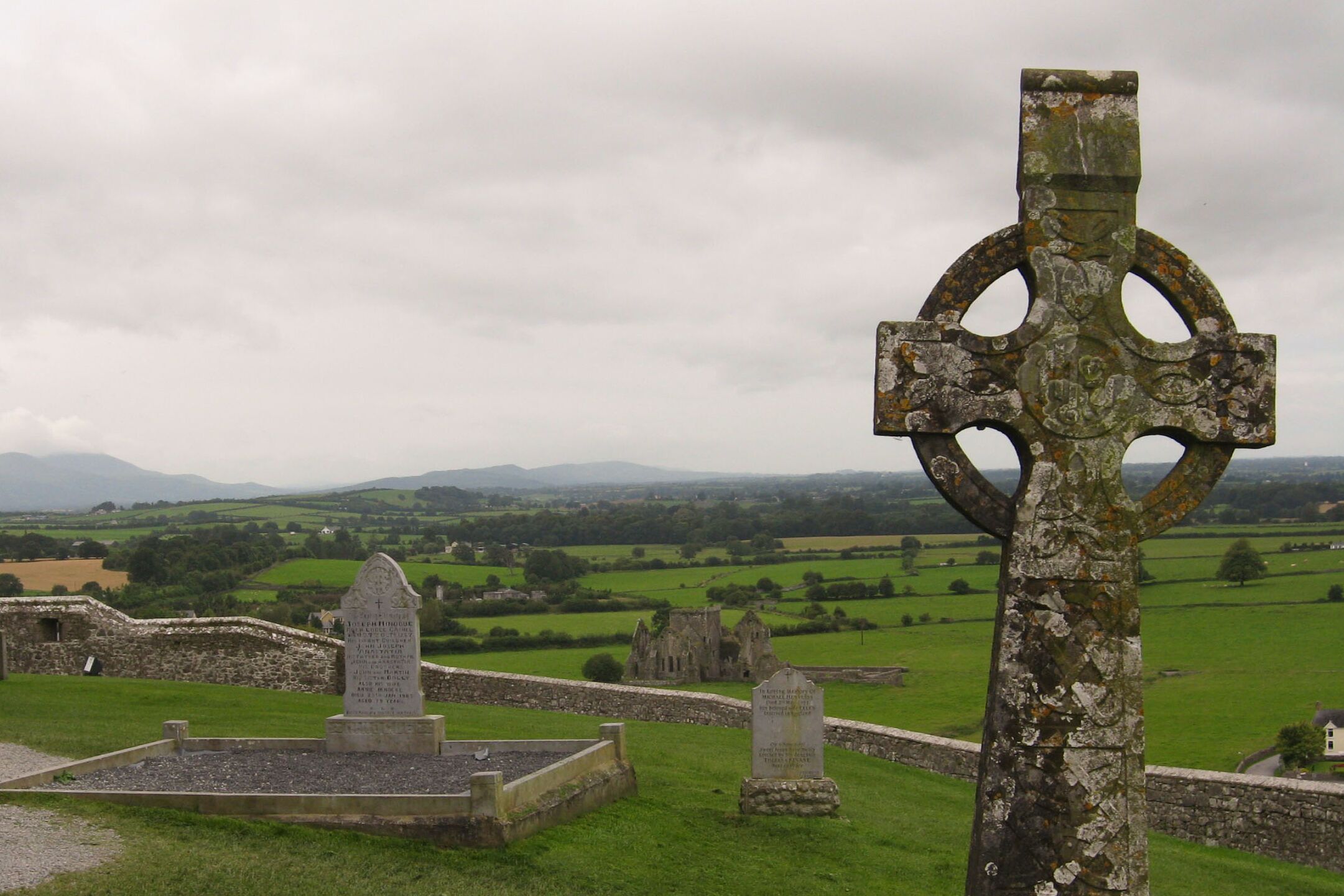 Keltisches Hochkreuz auf einem alten Friedhof in Irland, umgeben von Grabsteinen und grünen Wiesen.