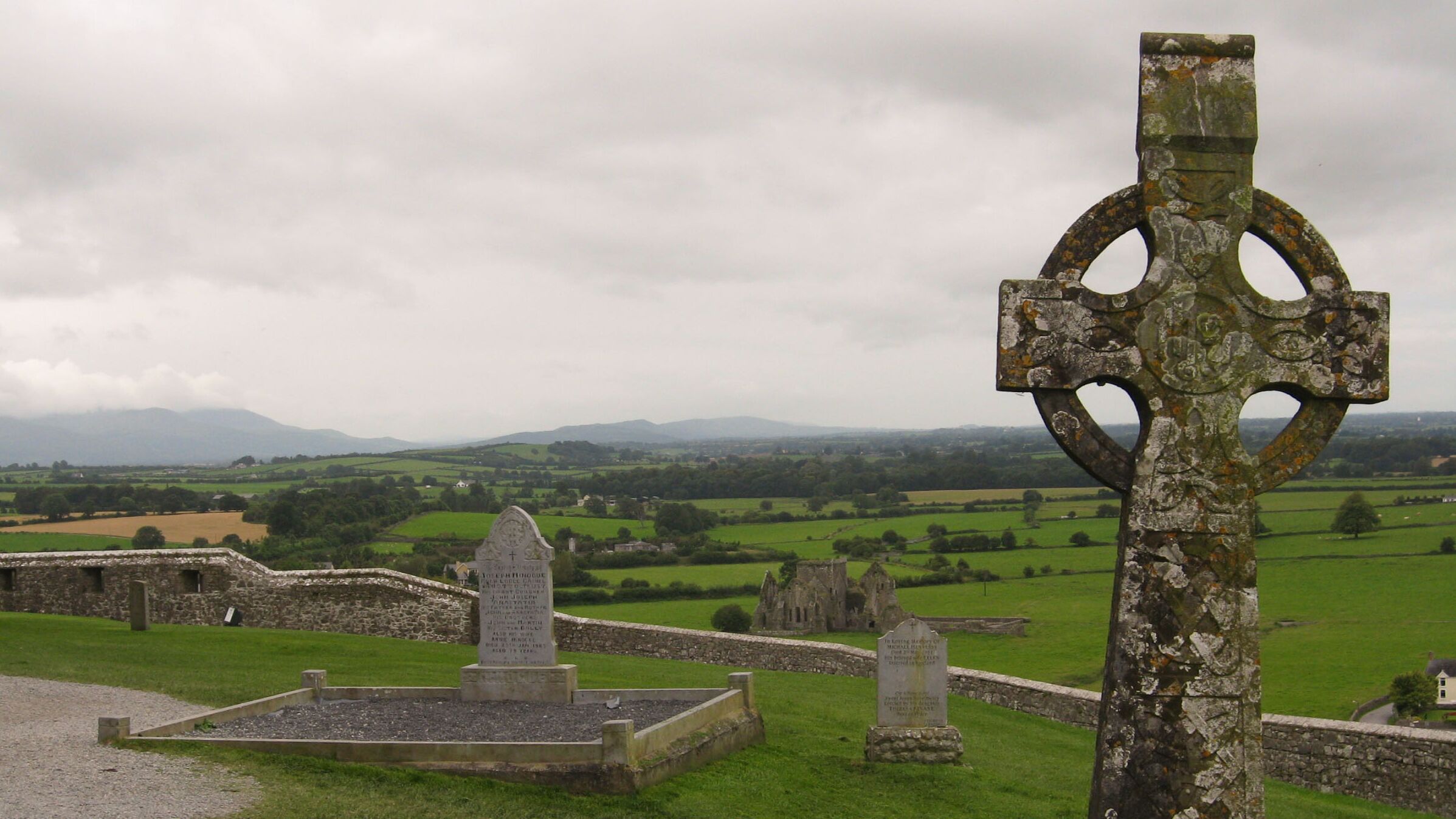 Keltisches Hochkreuz auf einem alten Friedhof in Irland, umgeben von Grabsteinen und grünen Wiesen.