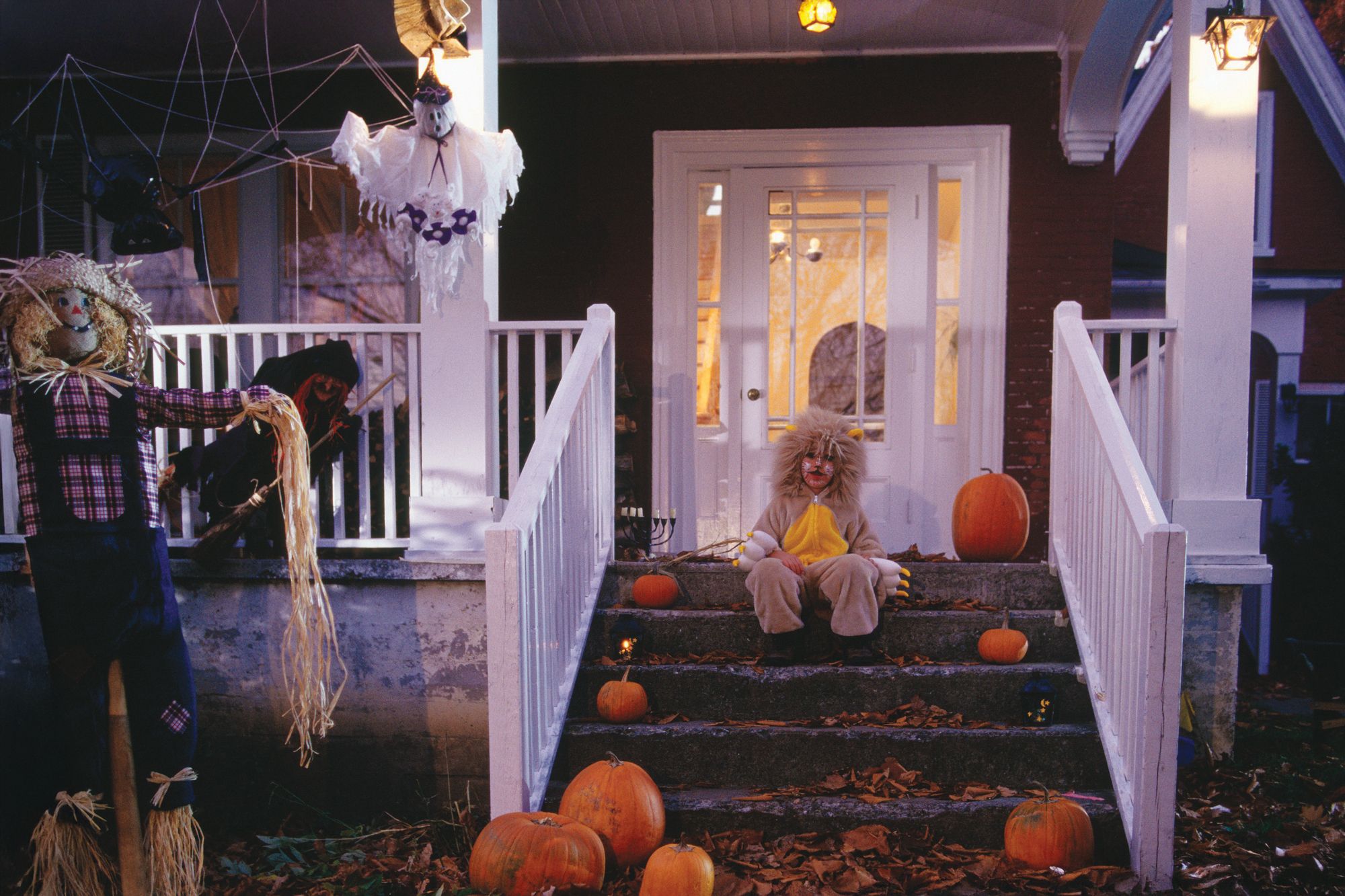 Ein kostümiertes Kind sitzt auf der Veranda eines mit Kürbissen und Spukgestalten geschmückten Hauses.