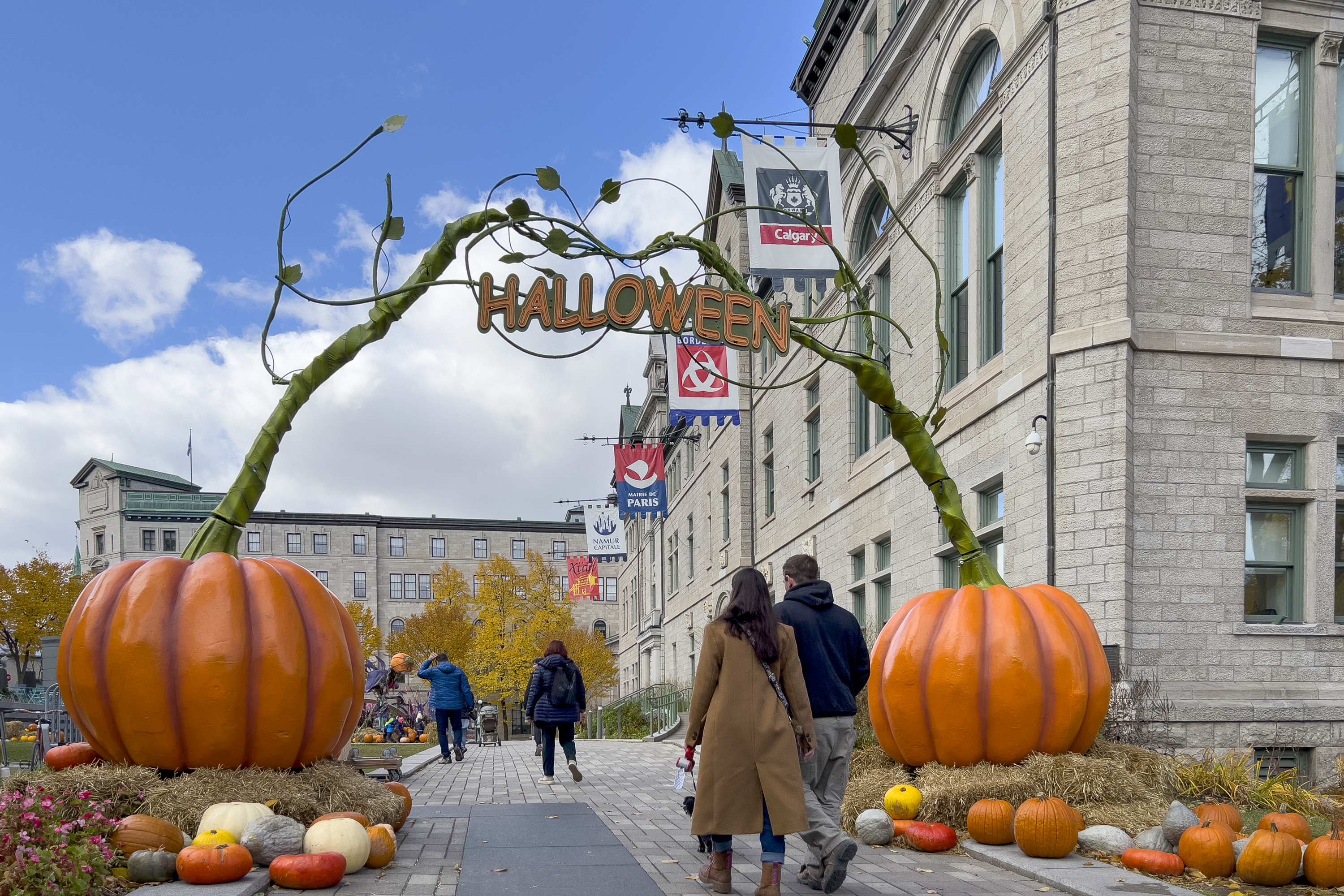 Zwei Fußgänger gehen unter einem dekorativen Bogen durch, auf dem der Schriftzug „Halloween“ prangt. Zwei riesige Kürbisse bilden die Ankerpunkte des Bogens.