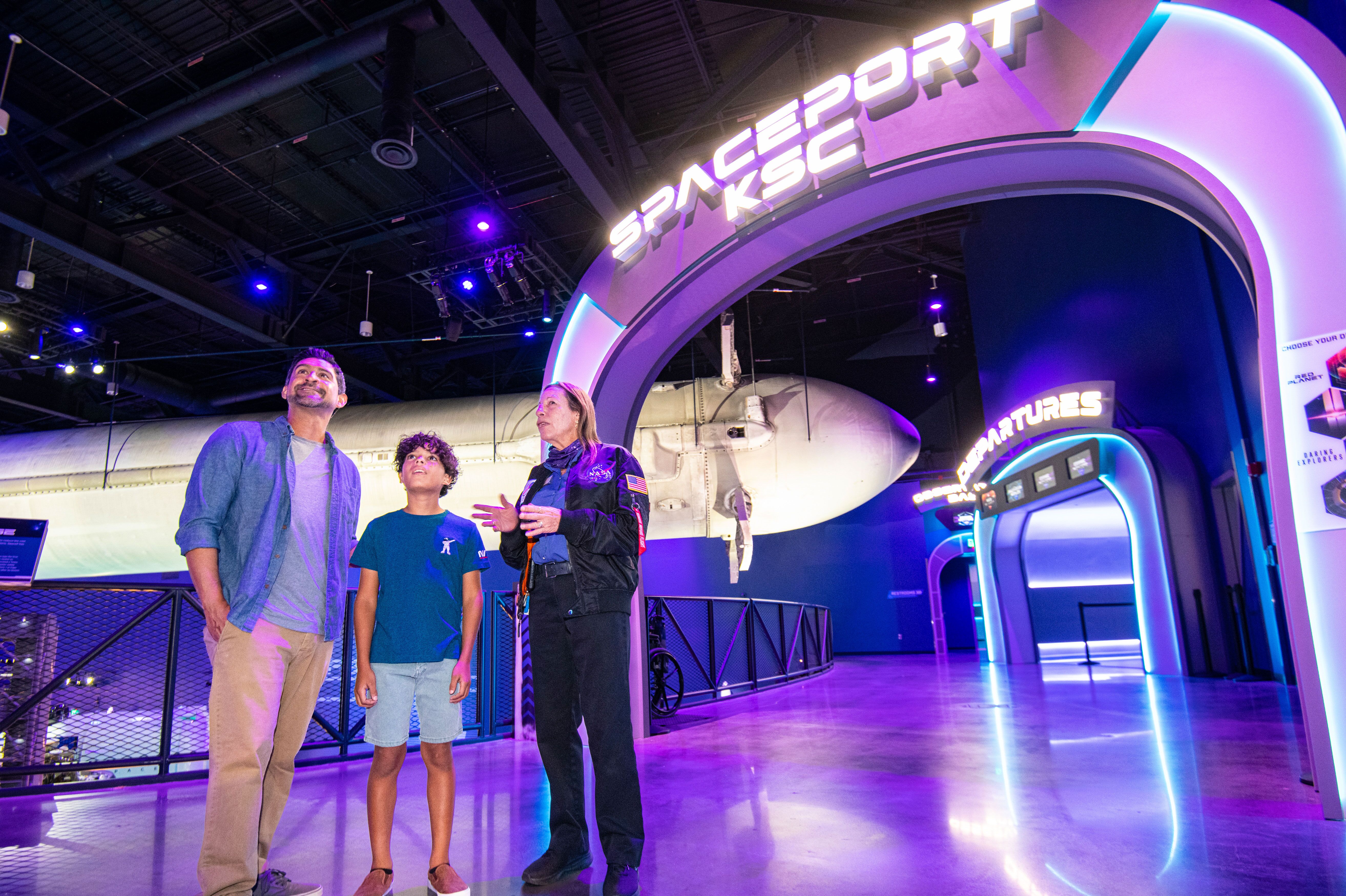 Mann und kleiner Junge unterhalten sich mit einem Mitarbeiter vor dem Eingang zum Spaceport KSC über die Ausstellungsstücke im Gateway im KSCVC