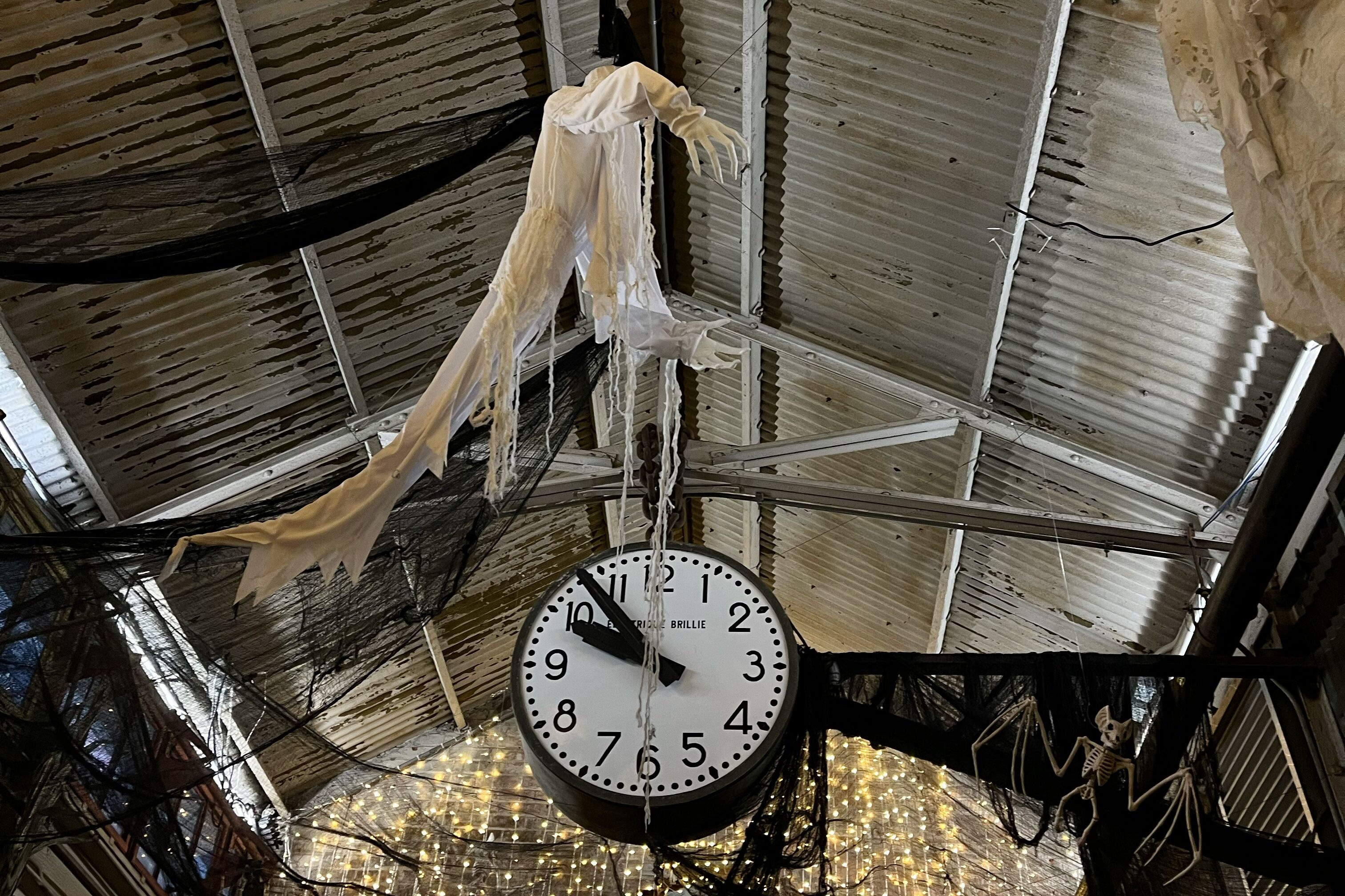An der Decke des Chelsea Market in New York hängt über einer großen Hallenuhr eine weiße Spukgestalt und schwarze Spinnweben.