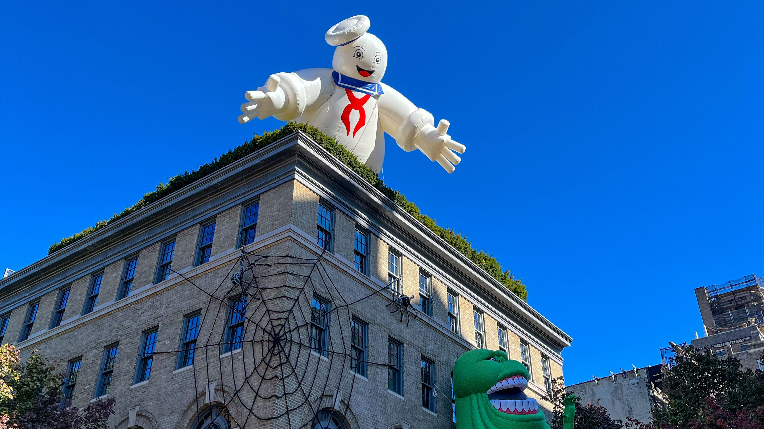Ein Gebäude in New York ist mit einem großen Spinnennetz geschmückt. An der Hausfassade klettert die Ghostbuster-Figur Schleim hoch, während über dem Dach übergroß der Marshmallow Man erscheint.