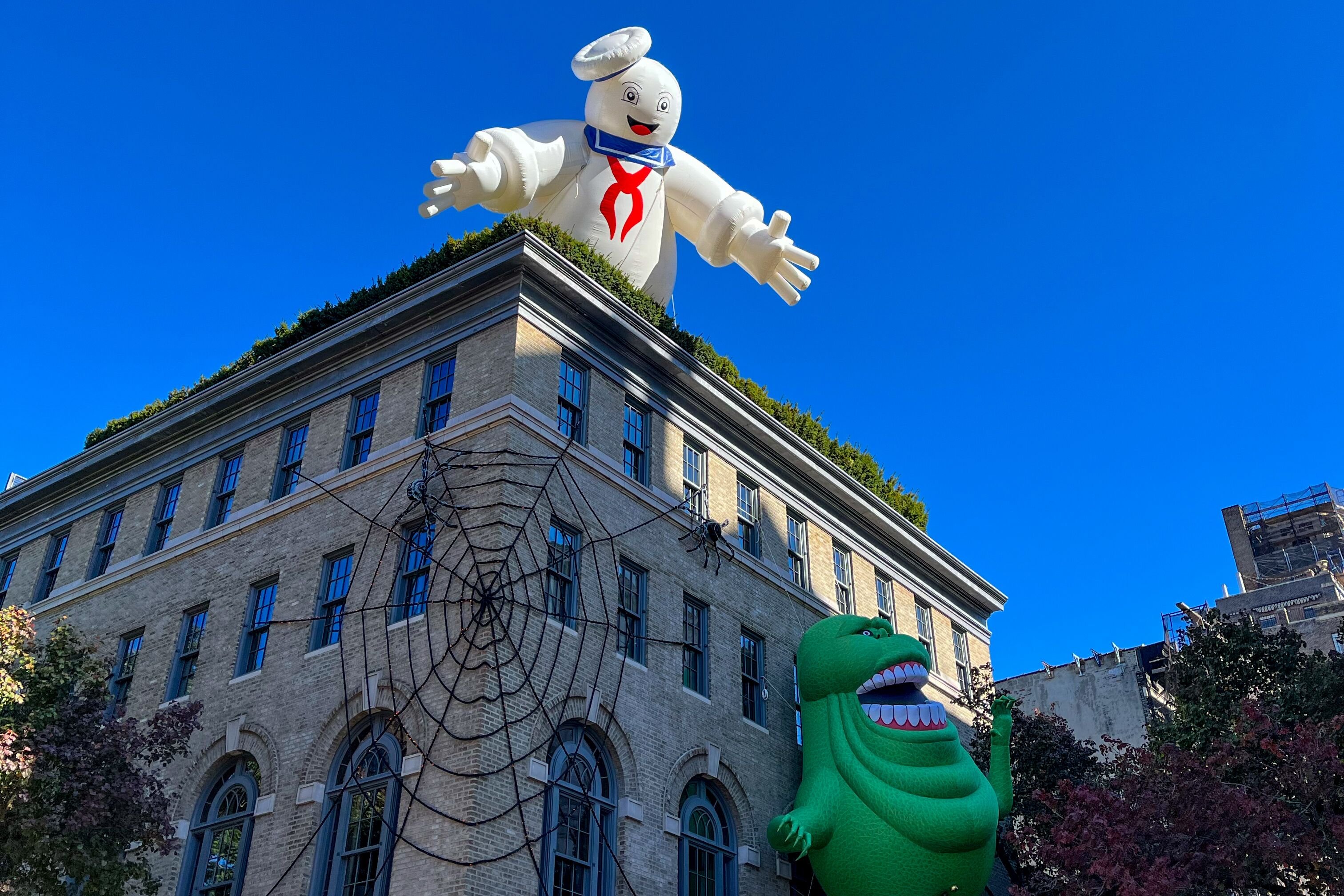 Ein Gebäude in New York ist mit einem großen Spinnennetz geschmückt. An der Hausfassade klettert die Ghostbuster-Figur Schleim hoch, während über dem Dach übergroß der Marshmallow Man erscheint.