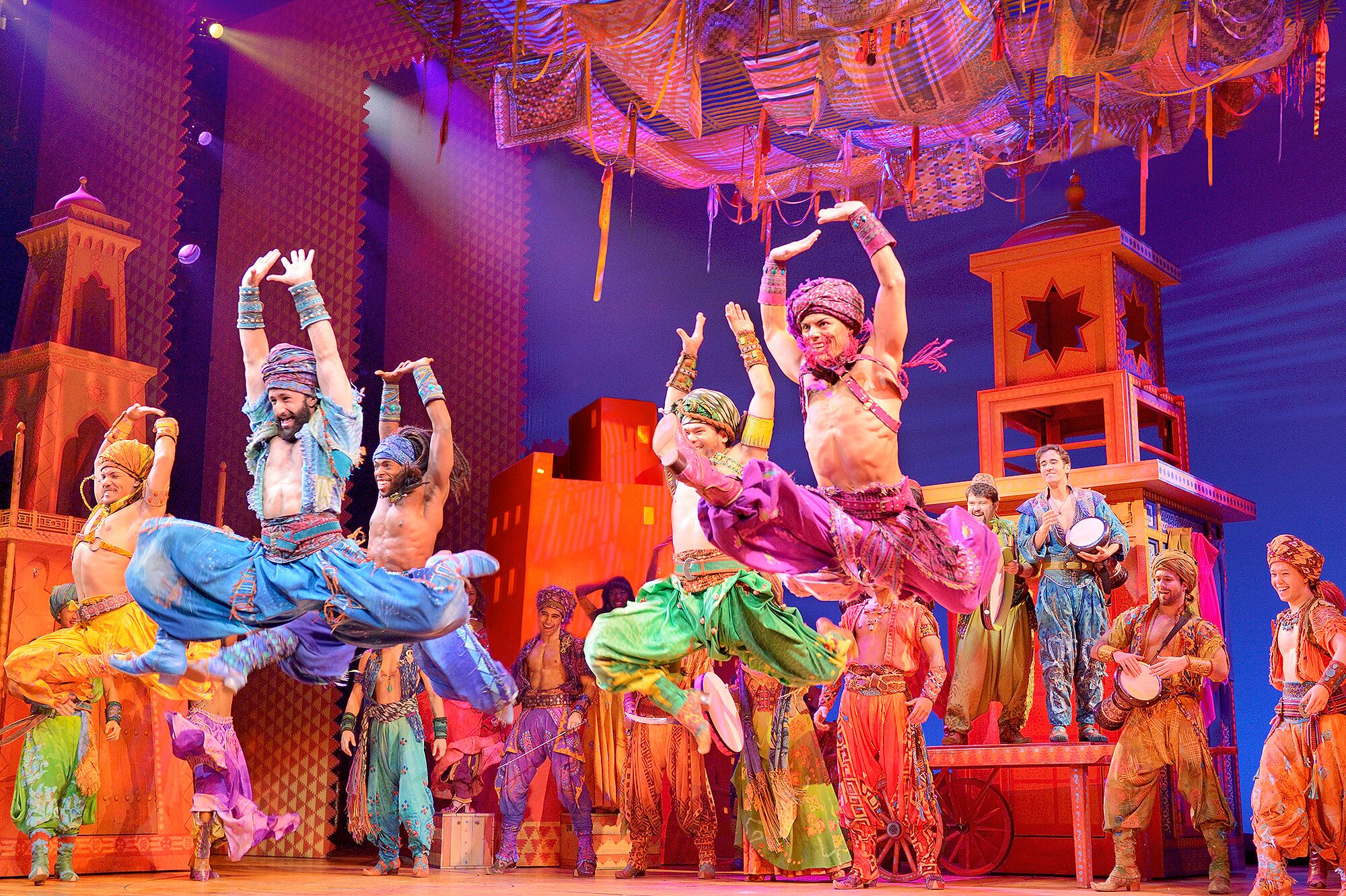 Tänzer aus dem Musical Aladdin in farbenfrohen Kostümen bei einer akrobatischen Performance auf einer mit orientalischen Elementen gestalteten Bühne.